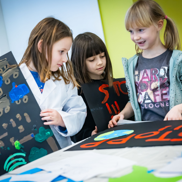 Auf dem Bild sind drei Kinder zu sehen, die gemeinsam um einen Tisch stehen und selbst bemalte Plakate vor sich liegen haben.