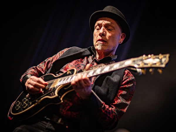 Musiker mit Hut spielt E-Gitarre auf dunkler Bühne.