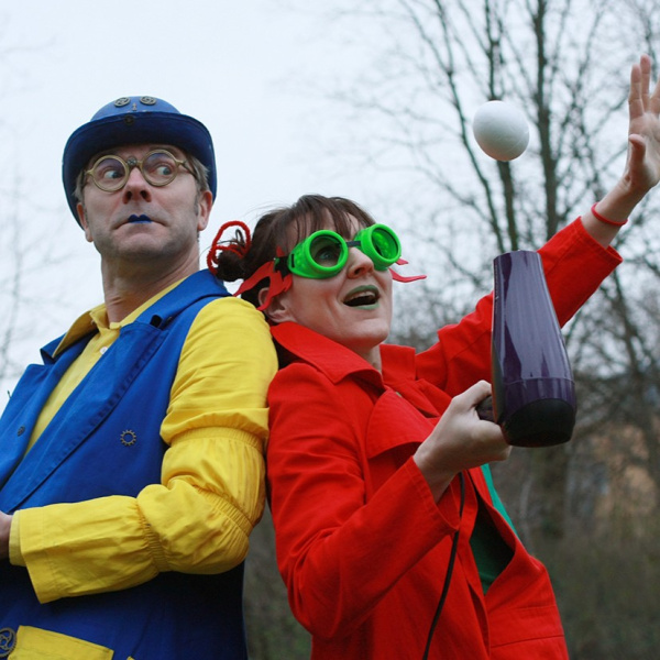 Zu sehen sind zwei Personen die bunt gekleidet sind und Rücken and Rücken zueinanderstehen. Links Helle und recht Leum. Helle blickt über die Schulter zu Leum, welche einen Föhn in der Hand hat und einen weißen Ball hoch fliegen lässt.