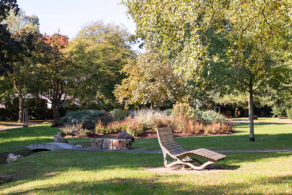 Blick in den Nicolaifriedhofspark mit Wiesenfläche und unterschiedlicher Bepflanzung sowie einer Parkbank