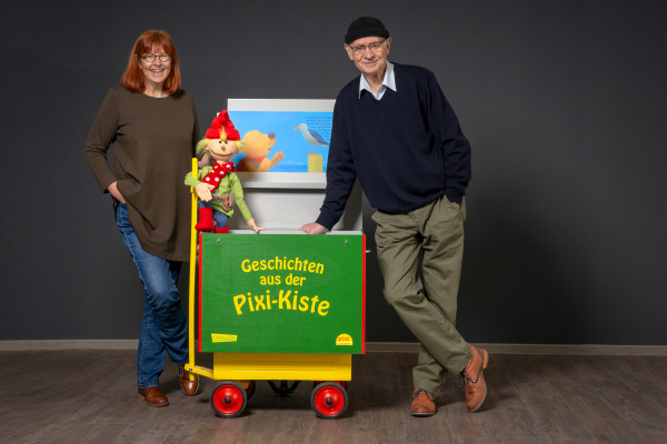 Darstellung von Herrn und Frau Paulsen an ihrer bunten Lesekiste vor dunklem Hintergrund.