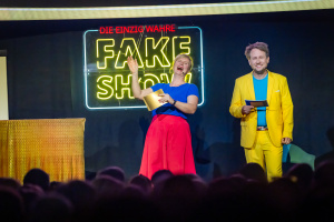 Zwei Personen auf einer Bühne mit "FAKE-SHOW"-Schriftzug im Hintergrund.
